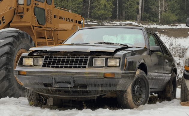 Grimy 1979 Ford Mustang parked on snow next to a yellow John Deere loader.