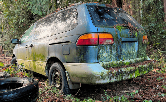 2000 Toyota Sienna minivan covered in green moss, sitting among trees after years parked.