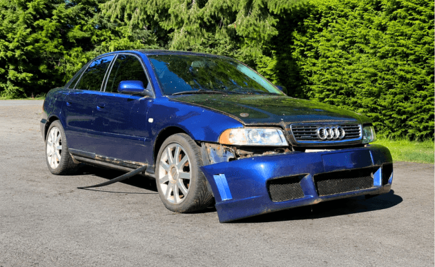 Blue 2001 Audi A4 B5 sedan with a battered front bumper, parked on gravel in front of a tall hedge.