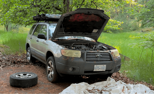 2006 Subaru Forester with its hood up in a green clearing, a spare wheel lying on the ground.