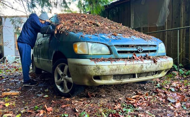Artur Stone leaning against an abandoned Toyota Sienna buried under fallen leaves beside a shed.