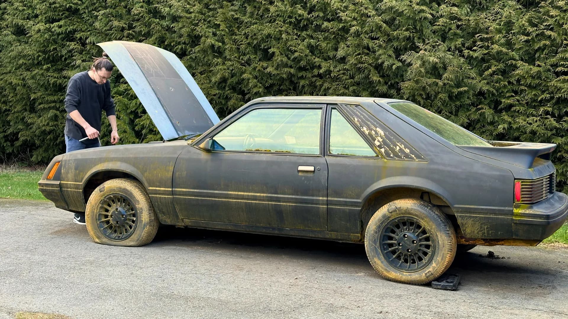 Artur Stone leaning under the open hood of a dust-covered black Ford Mustang, working on it.